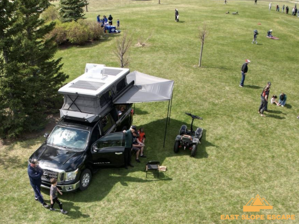 This is a drone shot of a camper. You can see the rooftop and extension of the camper and a few people gathered around, with more people in the field nearby.