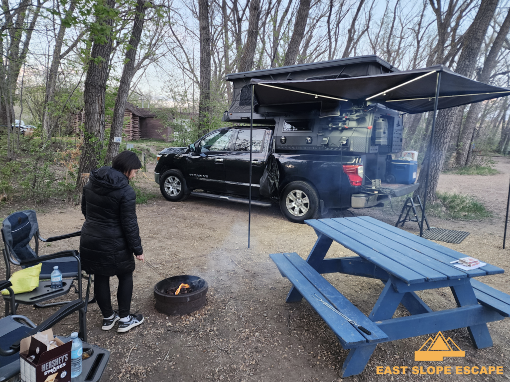 Woman stands in front of campfire with a camper in the background.